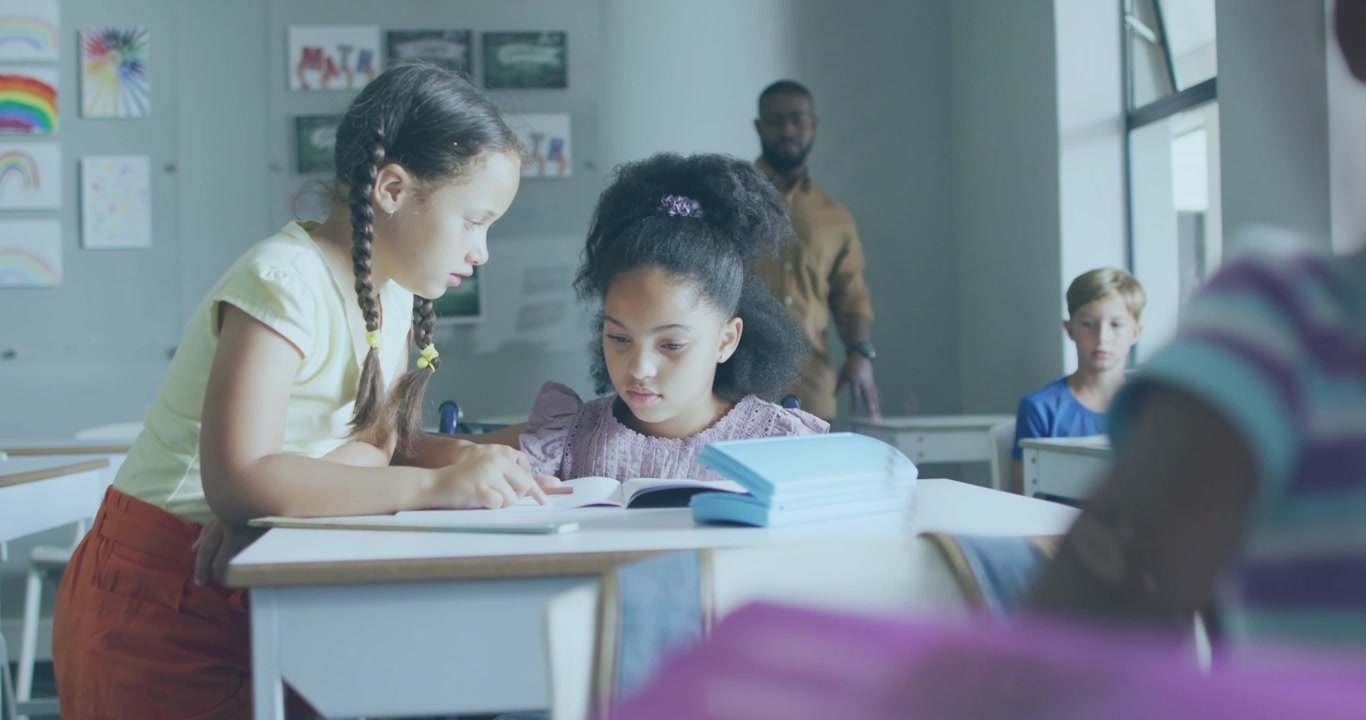Standing girl pointing at workbook, helping peer tracing with pencil, teacher watching classroom