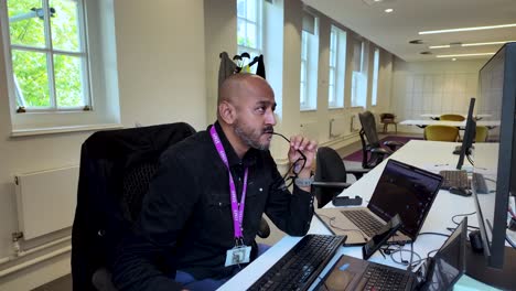 A-focused-individual-with-a-shaved-head-working-diligently-at-an-office-desk-with-two-monitors-and-a-laptop,-pensively-holding-glasses