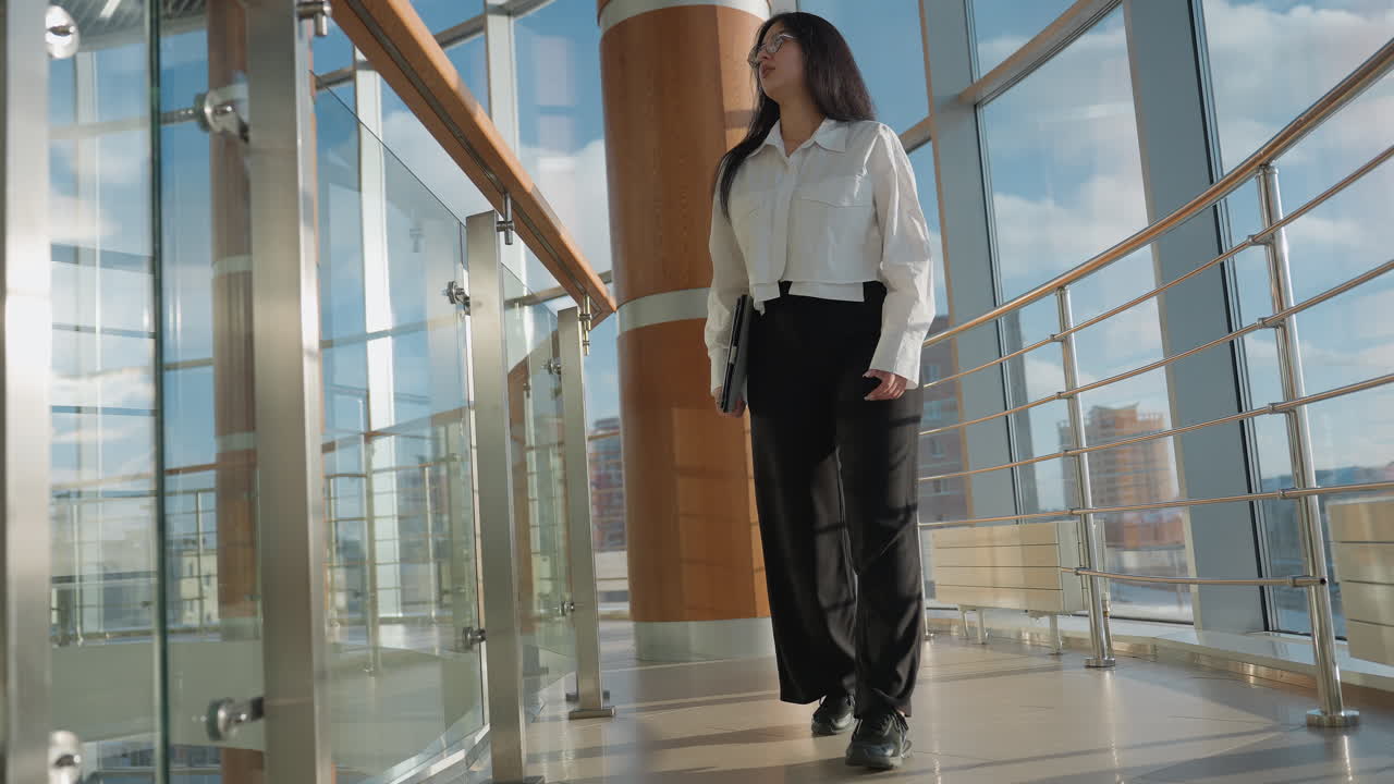 Woman in white blouse and black pants walks toward glass railing holding tablet in bright modern interior with large windows and wood paneled columns, glancing upward in thoughtful expression