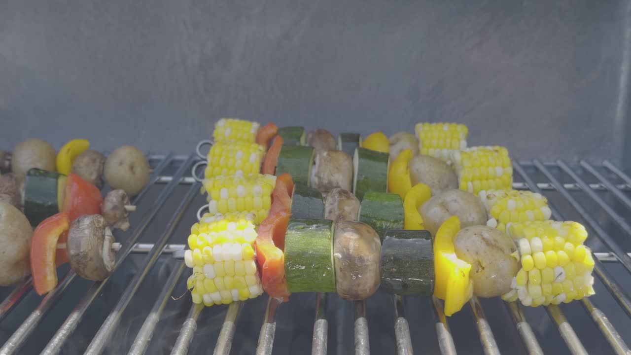 Closeup shot of vegetable skewers with corn, pepper, zucchini, potatoes and mushrooms are being cooked on a stainless steel bbq and brushed with oil. Smoke rises from the grill