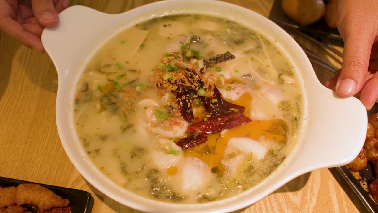 Hænder placerer en stor skål krydret Sichuan fiskesuppe med chiliolie på bordet.