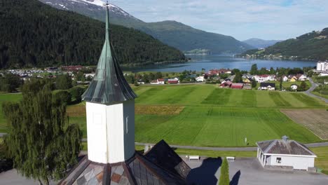 avance aéreo pasando muy cerca de la torre de la iglesia de loen - mostrando el edificio de la iglesia con vista panorámica sobre campos verdes y loenfjord en el fondo durante el verano - noruega