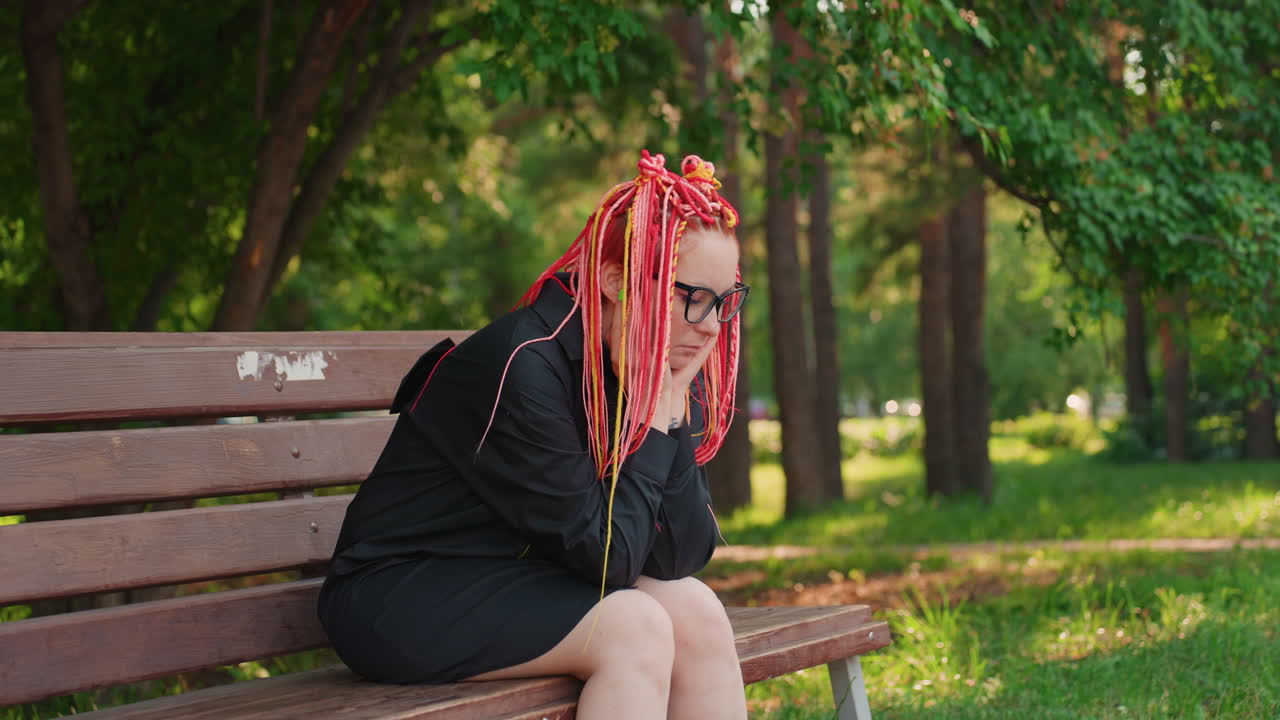 밝은 드레드락과 이어폰을 착용한 백인 여성이 벤치에 앉아 잎이 무성한 공원에서 옆모습을 보이고 있어요. 긴장한 자세와 먼 곳을 응시하는 모습, 여름 햇살과 조용한 도시의 녹지, 외로운 기다림의 풍경이 느껴져요.