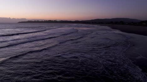 Pull-back-drone-shot-flying-directly-over-a-beach-with-waves-crashing-in-at-sunset