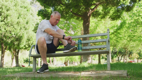un hombre mayor atando los cordones de sus zapatos mientras está sentado en un banco en el parque