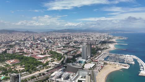 drone captures sweeping aerial view of coastal barcelona with barceloneta beach, port olimpic marina, tall modern building, cityscape, and distant mountain under blue sky on sunny day in spain