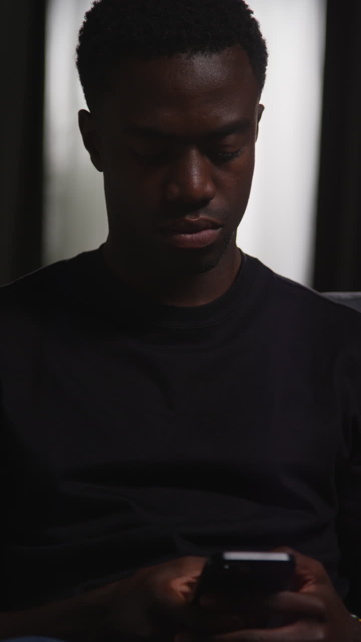 video vertical de primer plano de un joven infeliz y deprimido sentado en el sofá en casa enviando mensajes o navegando en el teléfono móvil