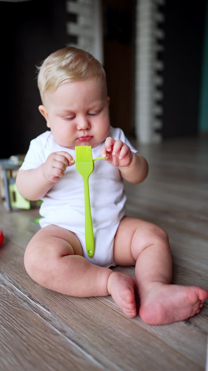 Beautiful plump Caucasian baby sits on the floor at home. Little kid in white bodysuit holds a kitchen brush. Vertical video.