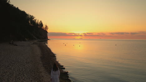 Vista-Trasera-De-Una-Joven-Recortada-Con-Vestido-Blanco-De-Verano-Caminando-Por-La-Costa-De-La-Playa-A-La-Hora-Dorada-Con-Un-Colorido-Disco-Solar-Sobre-El-Mar,-Seguimiento-Aéreo,-Espacio-De-Copia-En-Cámara-Lenta