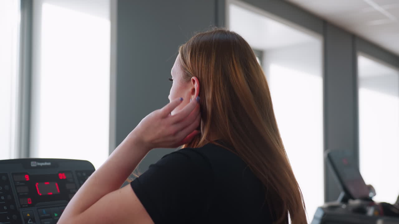 back view of young woman adjusting long hair while walking on treadmill in gym as another person leaves facility and man works out on adjacent machine with bright daylight through large windows