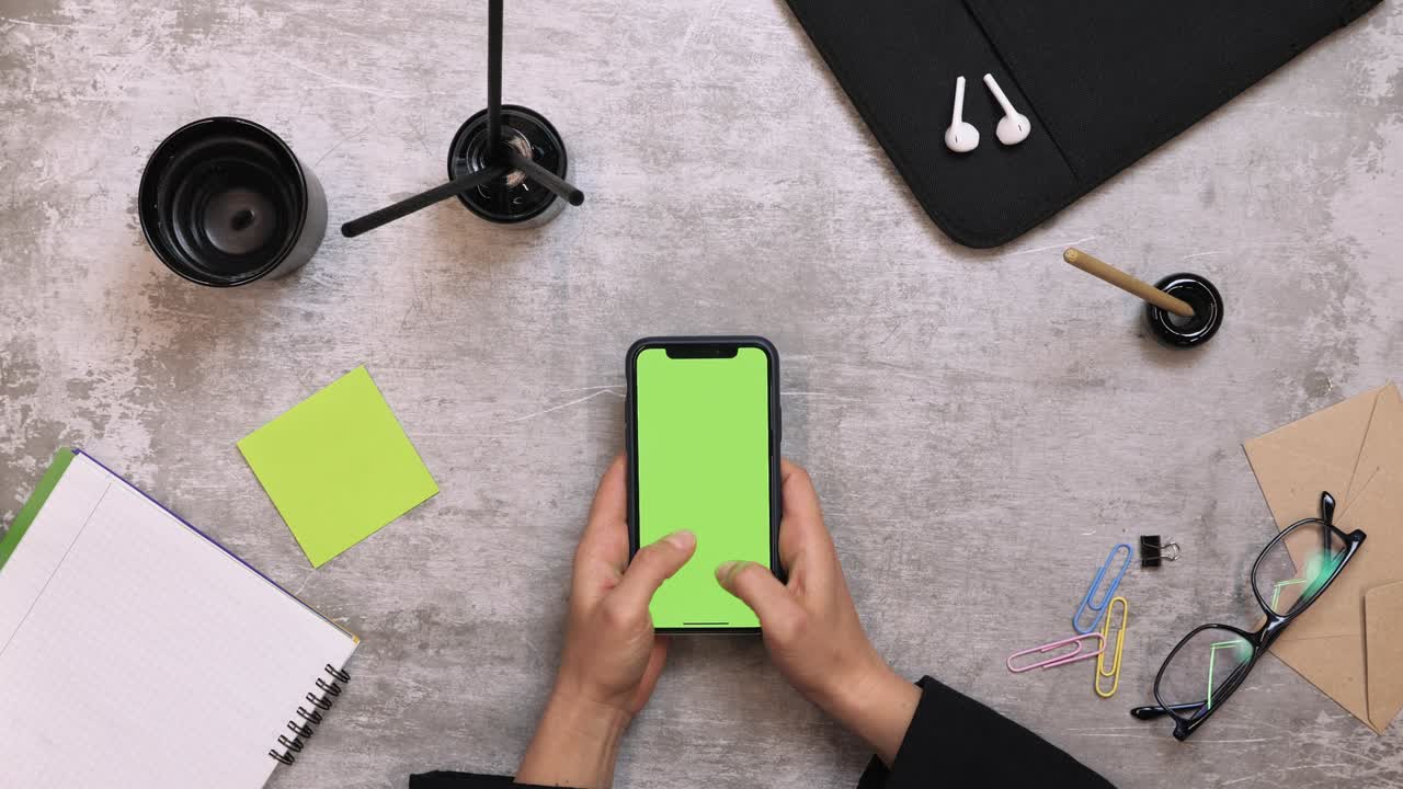 Businesswoman using smartphone, typing messages, letters while working at desk in office. Smartphone with green screen on work table, view from above. Woman using smartphone displaying chromakey