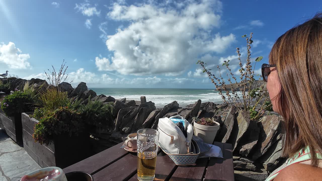 A person overlooks Trebarwith Strand Beach's vast sands and azure waves under a bright, sunny sky. Rocky foreground, sea stacks, and fluffy clouds add depth and drama