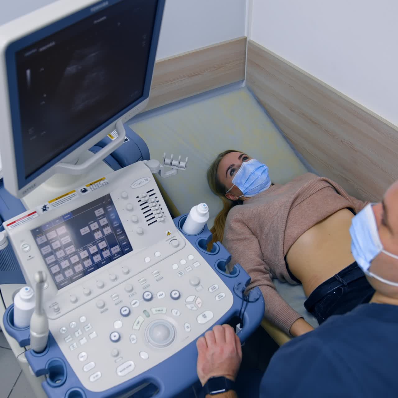 Woman in mask, sweater and jeans lies on the doctor's couch. Male medic puts ultrasonic device on patient's belly side. Top view