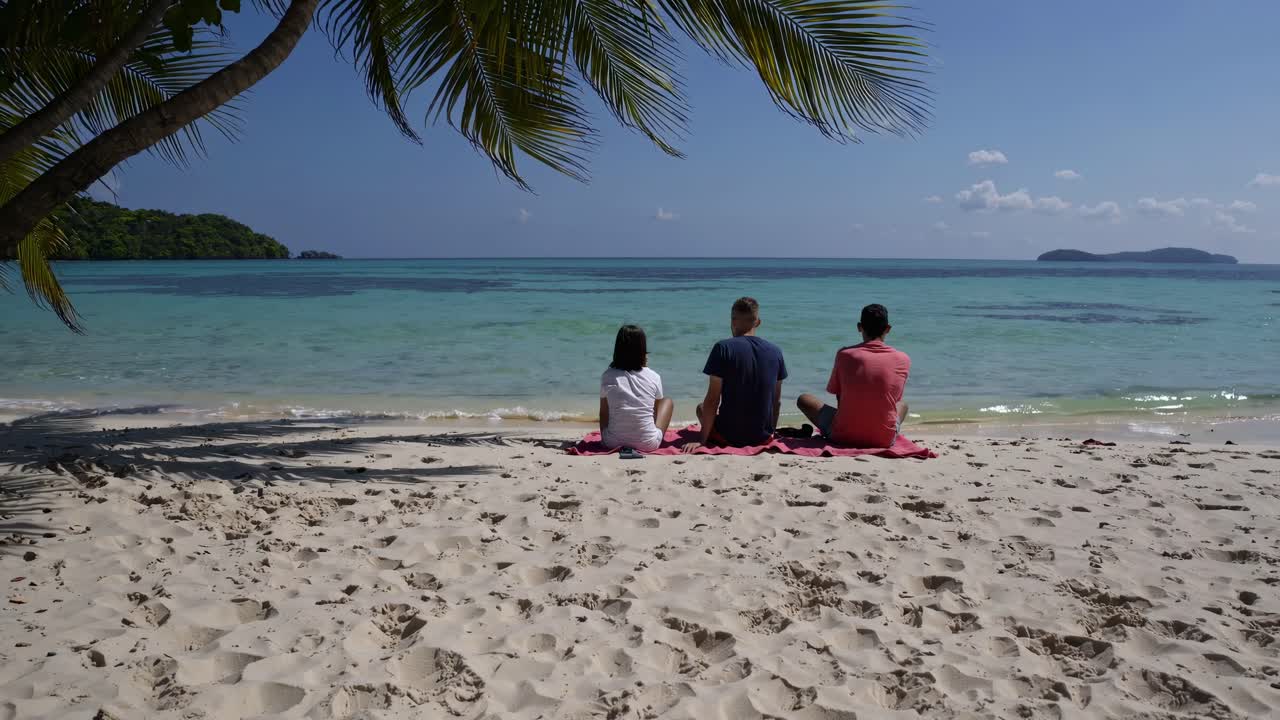 A serene beach video captures three people from behind, sitting on sand under palm trees
