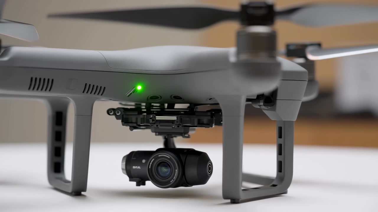 Düz bir yüzeyde duran tüketici quadcopter'ının alçak açılı ön üç çeyrek görünümü, yanan yeşil durum ışığı, asılı kamera gimbalı, pervane kolları, iniş takımları ve temiz stüdyo ortamını gösteriyor.