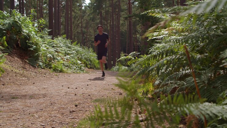 Low Angle Shot Of Man Exercising Doing Work Out Outdoors Running Along Track Through Forest Towards Camera Wearing Sports Clothing Shot In Real Time