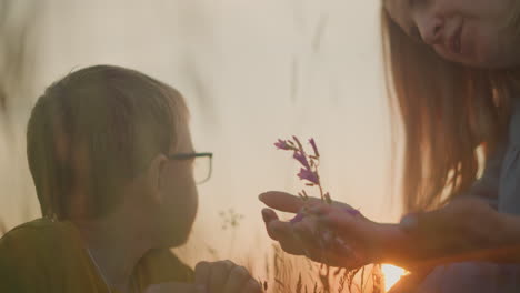 a tender moment as a woman in a blue dress offers a purple flower to a young boy wearing glasses, inviting him to smell it during a serene sunset, capturing a peaceful connection with nature