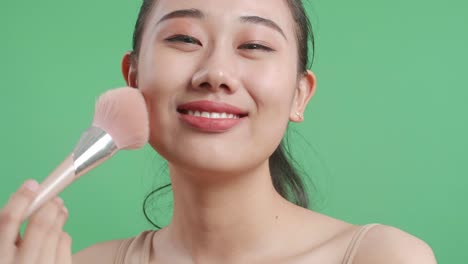 un primer plano de una hermosa mujer asiática agitando las mejillas y sosteniendo un pincel de maquillaje mientras sonríe a la cámara en el fondo de la pantalla verde en el estudio