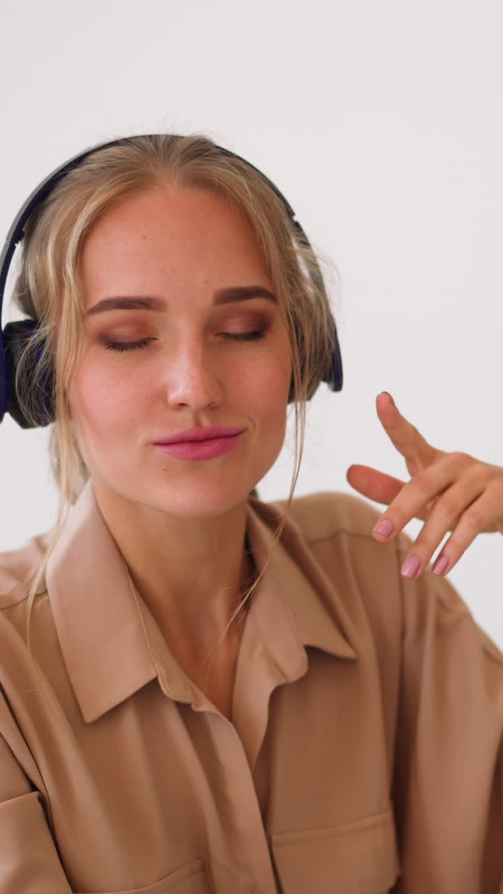 Blonde woman listens to music in headphones and makes dance moves with closed eyes sitting at workplace with contemporary laptop slow motion closeup