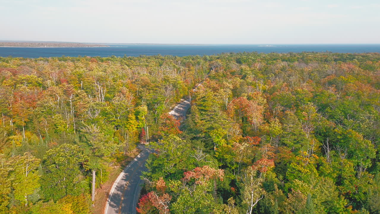 A narrow winding road snakes through a canopy of red, yellow, and green trees, drawing the eye toward a shimmering blue lake that meets the horizon in crisp autumn air