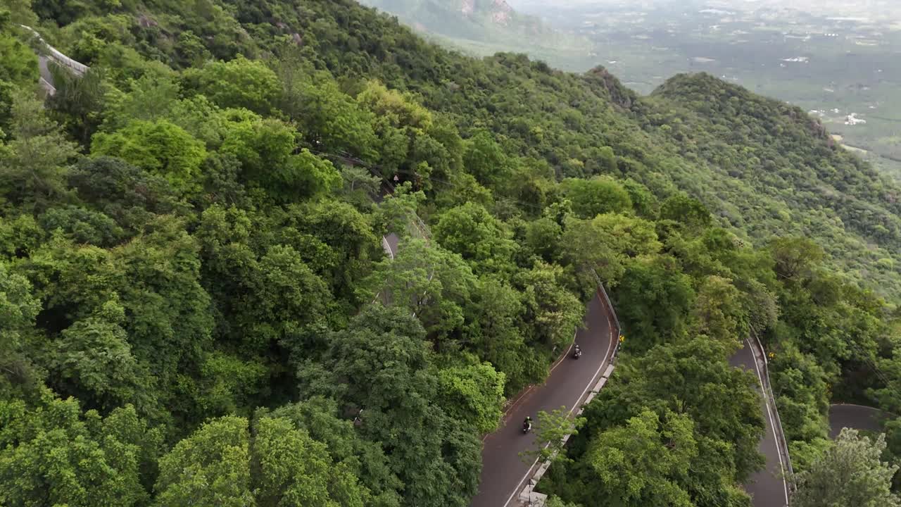 Scenic aerial footage of a winding road beginning its climb towards Kolli Hills, Tamil Nadu. Surrounded by rural fields and hilly dense forest slopes