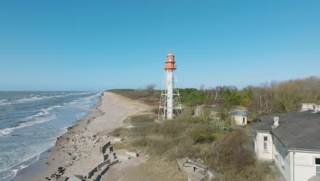 aerial establishing view of white colored pape lighthouse, baltic sea coastline, latvia, white sand beach, large waves crashing, sunny day with clouds, wide ascending drone shot moving forward