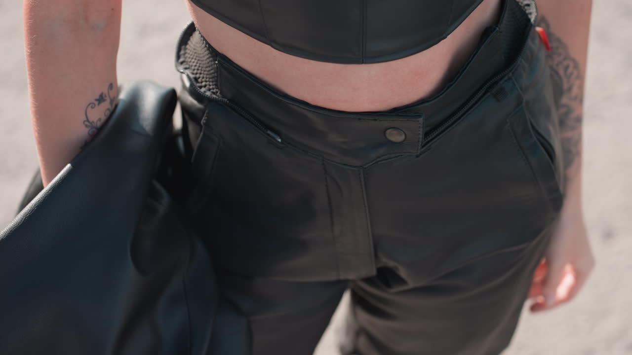 Close up of girl with tattooed hand holding dark sunglasses while wearing black leather crop top and chain necklace outdoors under sunlight, showing stylish details with soft blurred background