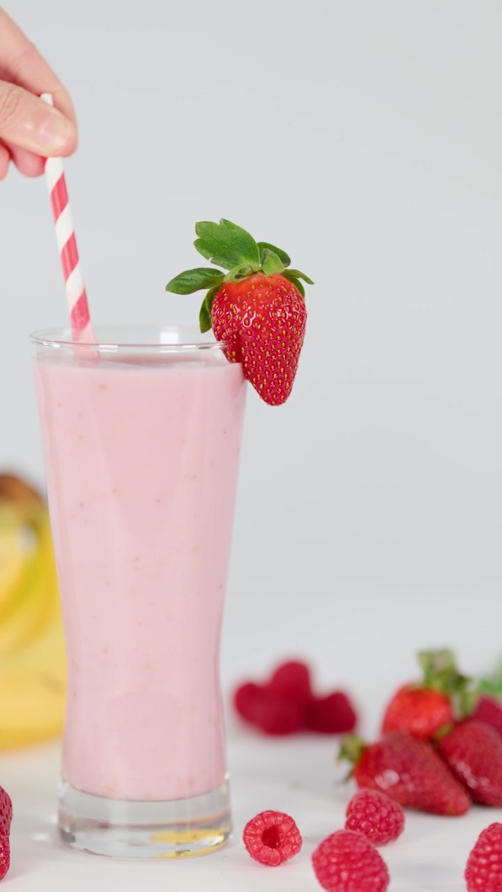 Una mano aggiunge una cannuccia a righe e una guarnizione di fragola fresca a un frullato rosa cremoso in uno studio luminoso.