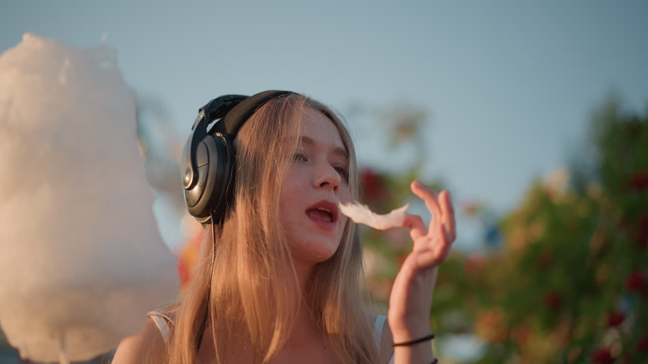 Giovane donna bianca che assaggia zucchero filato con le cuffie, primo piano intimo di luna park, occhi chiusi in un sereno momento ASMR, assaporando lentamente lo zucchero, bokeh colorato delle giostre sullo sfondo, atmosfera da influencer
