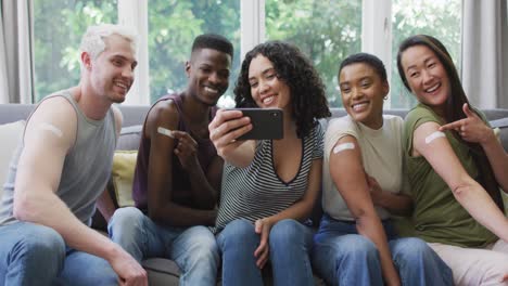 grupo de jóvenes diversos mostrando sus hombros vacunados y tomando una selfie en casa