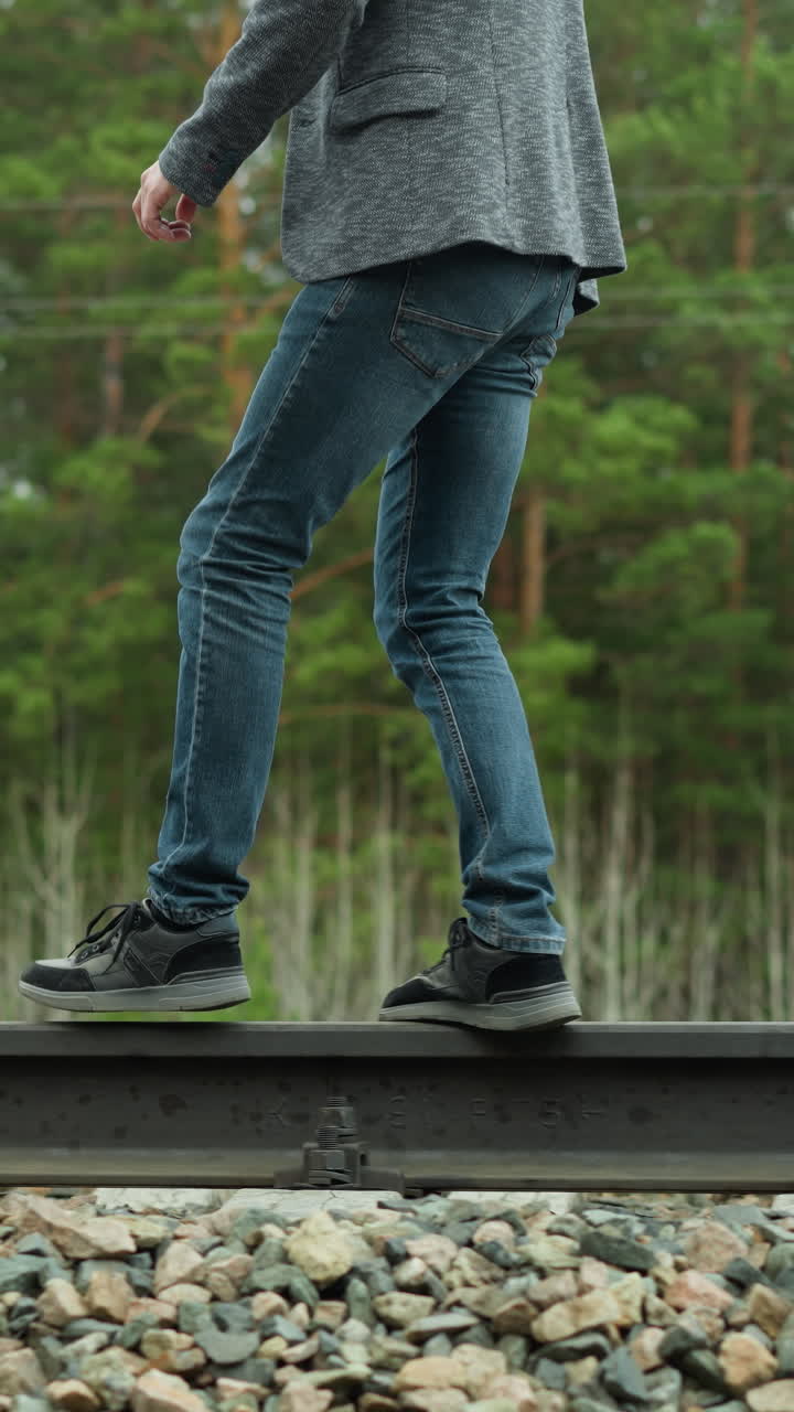철도 트랙을 따라 걸어가는 파란 청바지와 검은 스니커를 입은 사람의 클로즈업 뷰, 조심스럽게 균형을 잡고, 배경에는 울창한 초록색 나무가 있습니다.