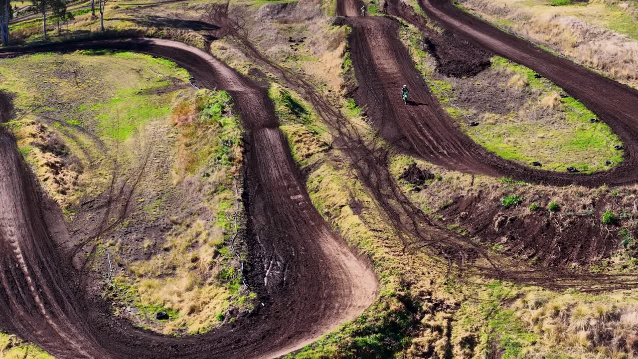 A motocross rider navigates a dirt track curve, launching into the air. Captured by drone in bright daylight, surrounded by bushland and eucalyptus trees
