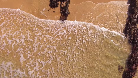 Aerial-view-looking-down-on-to-small-ocean-waves-lapping-onto-a-sandy-beach-with-some-rocks,-while-the-camera-slowly-descends-in-the-Gower-in-Wales