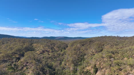 Aerial-rises-above-the-Barimoon-area,-and-forest-covered-hills-to-expose-the-Boyd-Burnett-Rail-Trail-cutting-through-the-woodlands-with-views-to-Kalpower-State-Forest-Central-Queensland-Australia