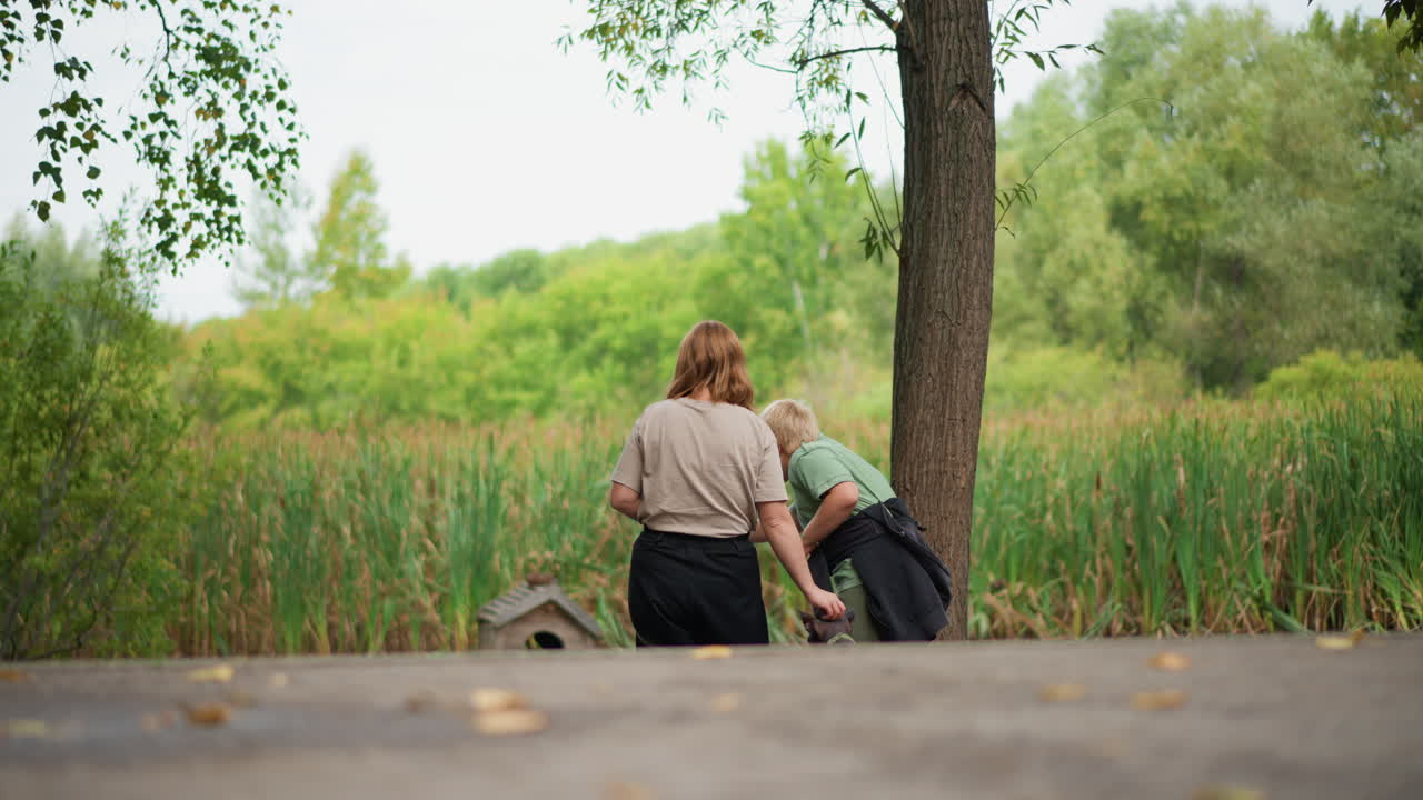 Woman And Boy By Water, Reflective Moment As Figures Observe Still Lake Surroundings In Summer, Gentle Scene Showing Woman And Boy Seated Peacefully Overlooking Calm Water And Tall Reeds