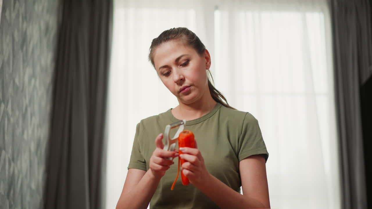 Focused woman in green t-shirt peeling carrot with handheld peeler in softly lit room, natural light shines through sheer curtains as she carefully removes carrot skin with concentrated expression