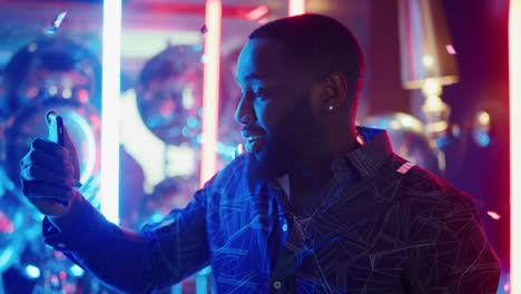 Smiling-afro-man-talking-on-video-on-neon-background.-Guy-holding-mobile-in-club