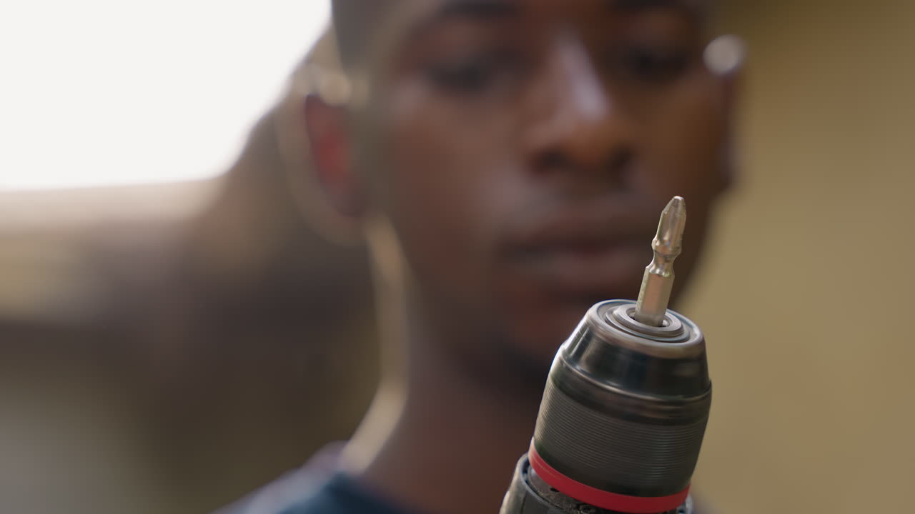 Black apprentice carpenter inspects drill closeup, focused young black man in blurred workshop background, power drill held steady with metal bit glinting, concentrated expression, carpentry training
