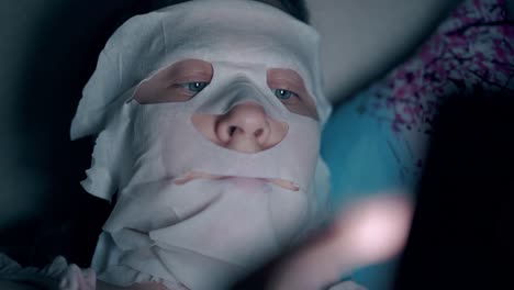 person in sheet mask lies on blue pillow with flowers