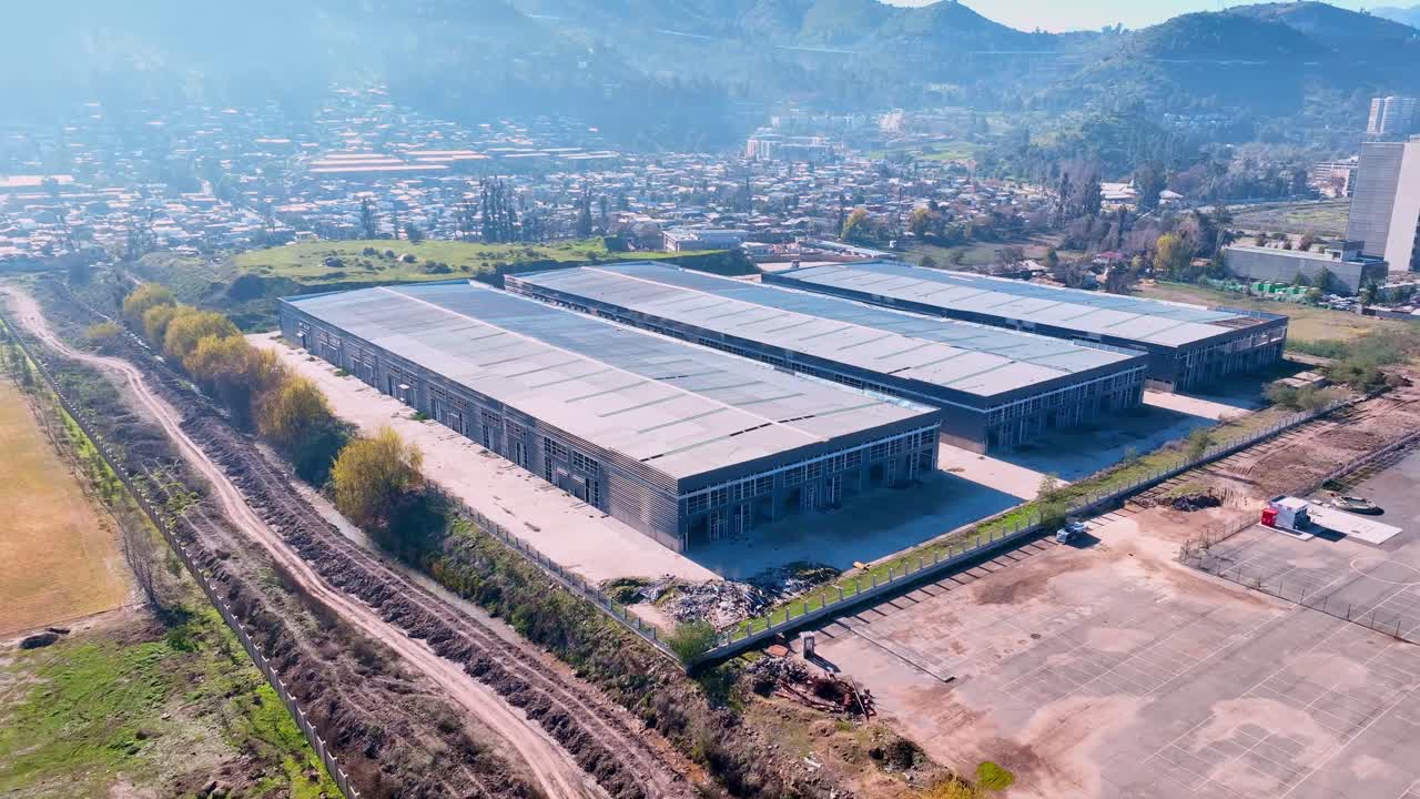 Orbiting drone aerial of large modern warehouse with metal roof, industrial facility surrounded by mountains and urban landscape, Chile