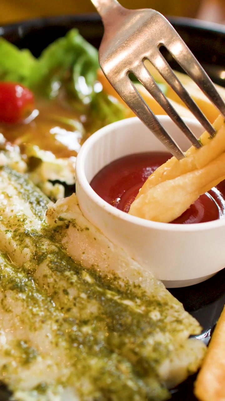 Una forchetta intinge una patatina fritta dorata nel ketchup rosso accanto a pesce alla griglia e pesto.