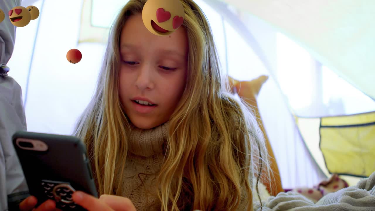 Girl lying blanket tent tapping smartphone screen causing swirling emoji swarm for tech exploration