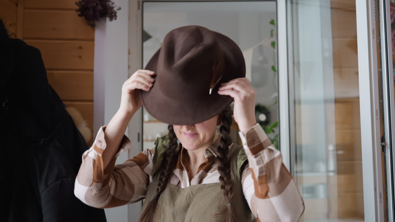 Window view shows female farmer walking to corner where jacket and hat hang on hook then picking hat and placing on head inside wooden room under natural light under warm indoor setting