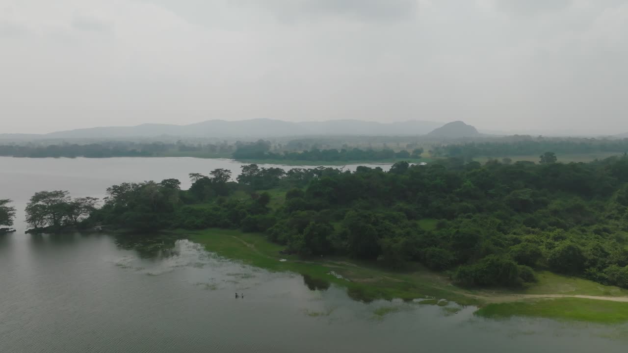 Aerial view of wetlands and forests along the shoreline of a lake in Sri Lanka, framed by misty mountains in the background