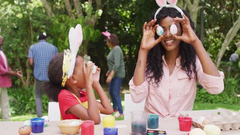 Animation-of-happy-african-american-mother-and-daughter-painting-easter-eggs-outdoors,-having-fun
