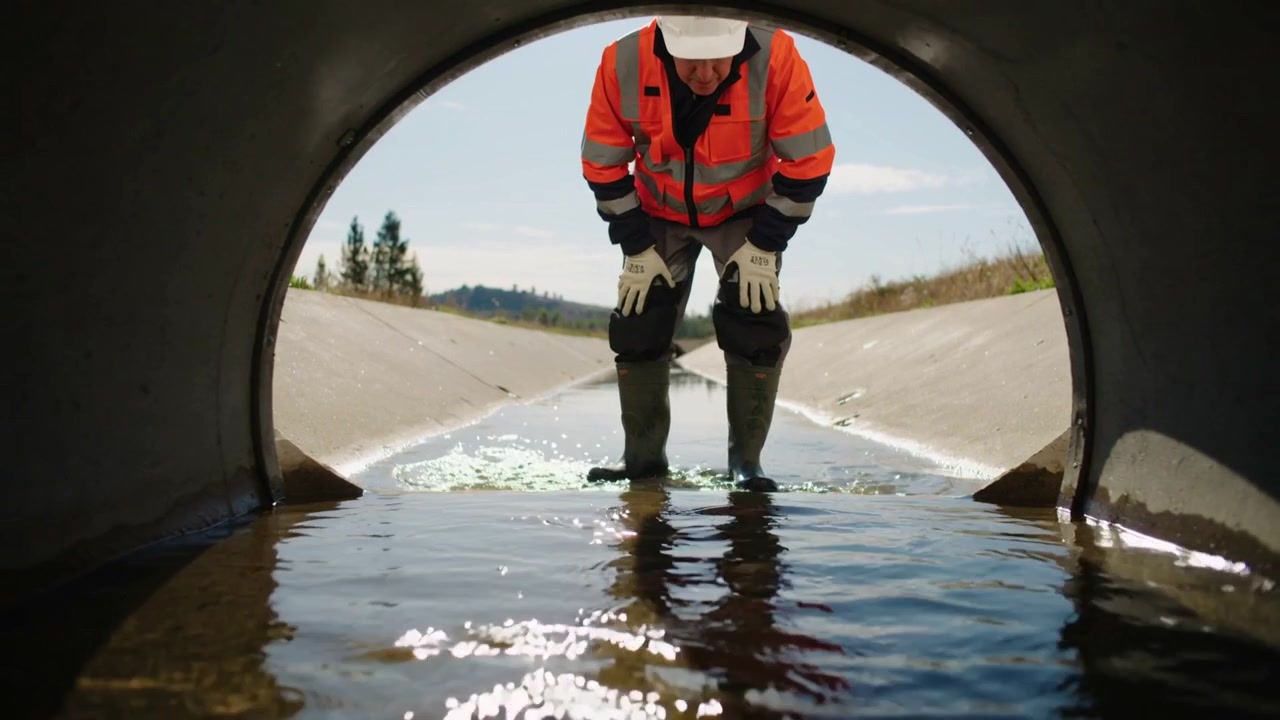 Inspecteur stapt in het water en inspecteert de betonnen duikeruitlaat langs de weg, gekleed in een oranje jack.