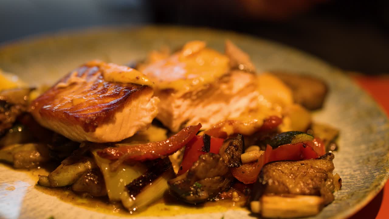 Hand slices grilled salmon fillet atop gourmet vegetables under warm, ambient restaurant lighting, close-up view