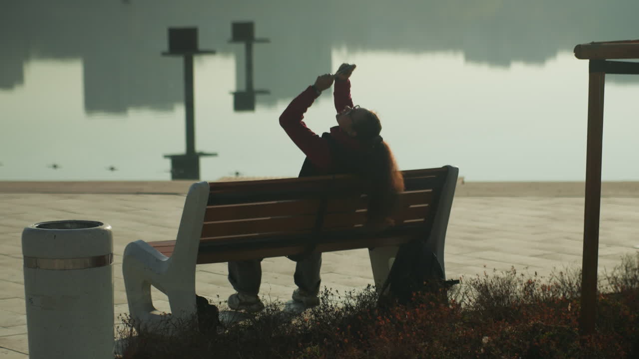 Person on bench by waterfront, phone in hand capturing selfie and playful gestures sunrise light highlights hair, city skyline reflected in water, content creator vibe, social connectivity, candid