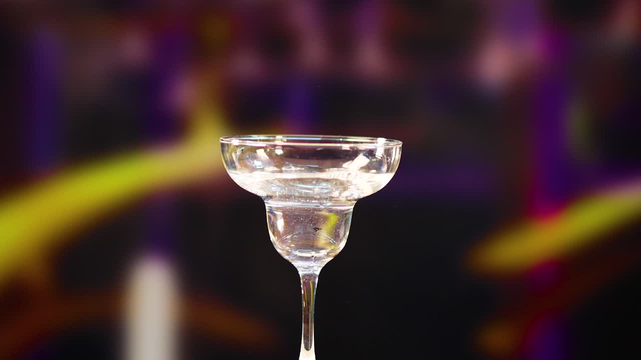 Un bicchiere da martini si riempie di liquido su uno sfondo colorato e sfocato, ripreso al rallentatore con illuminazione drammatica