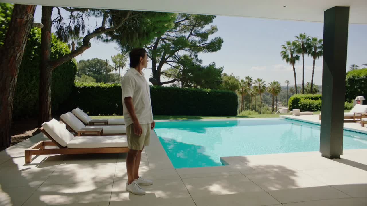 Noticing hillside on covered deck, man wearing shirt and shorts stepping toward pool, with lounges
